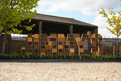 Large wooden lettering spelling 'JIMMY's' mounted outdoors in front of a rustic wooden pavilion, with gravel ground in the foreground and trees on both sides at Jimmy's Farm and Wildlife Park entrance.